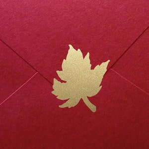 May include: A red envelope with a gold maple leaf emblem. The envelope is made of textured paper and has a classic triangular flap closure. The gold leaf is centered on the front.