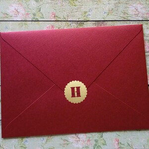 May include: A deep red envelope with a gold sticker bearing the letter "H". The envelope is sealed and has a textured finish. The background features a floral pattern on a wooden surface.