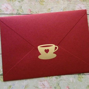 Könnte beinhalten: Ein roter Umschlag mit einem goldfarbenen Teetassen- und Herzdesign. Der Umschlag ist verschlossen und hat eine matte Oberfläche. Das Teetassen-Design ist mittig auf der Vorderseite des Umschlags platziert.