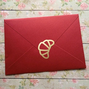 May include: A red envelope featuring a gold croissant emblem. The envelope is sealed and has a minimalist design. The background shows a floral pattern.
