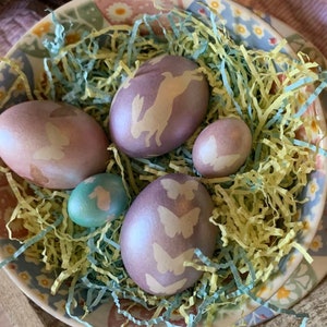 K&ouml;nnte beinhalten: Eine Sch&uuml;ssel gef&uuml;llt mit pastellfarbenen Ostereiern, die mit Hasen- und Schmetterlingsmotiven verziert sind. Die Eier liegen in zerfetzten gelben und blauen Papier.