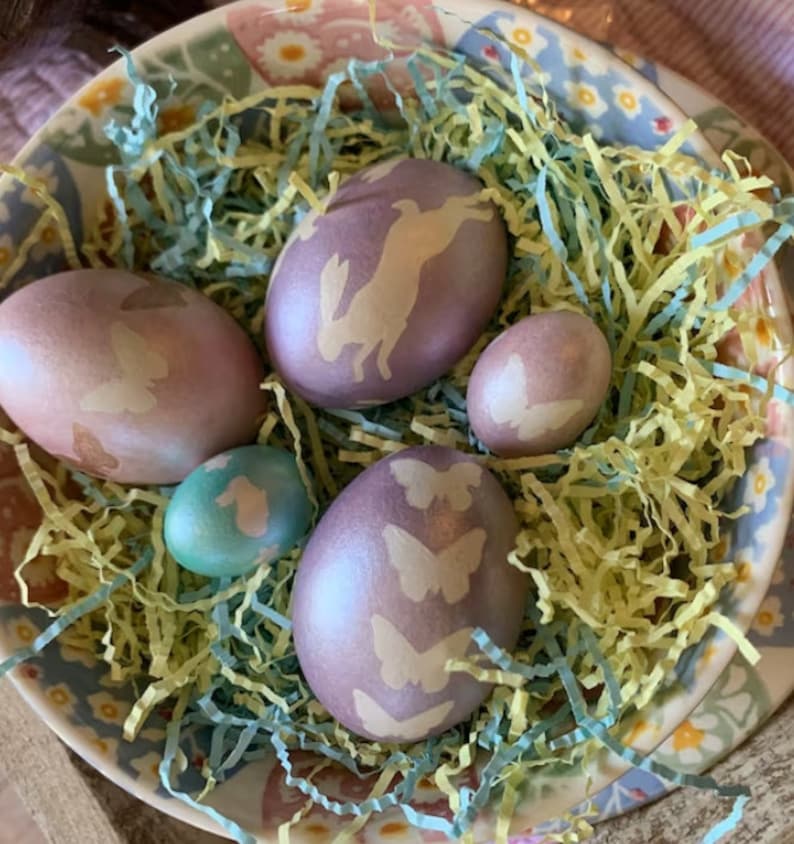 K&ouml;nnte beinhalten: Eine Sch&uuml;ssel gef&uuml;llt mit pastellfarbenen Ostereiern, die mit Hasen- und Schmetterlingsmotiven verziert sind. Die Eier liegen in zerfetzten gelben und blauen Papier.
