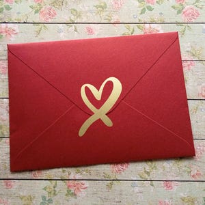 May include: A red envelope with a gold heart design. The envelope is closed and has a simple, elegant design. The heart is a stylized, cursive shape.