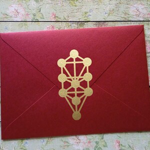 May include: A red envelope with a gold Kabbalistic Tree of Life symbol. The envelope is made of textured paper and has a classic envelope shape. The gold symbol is centered on the front.