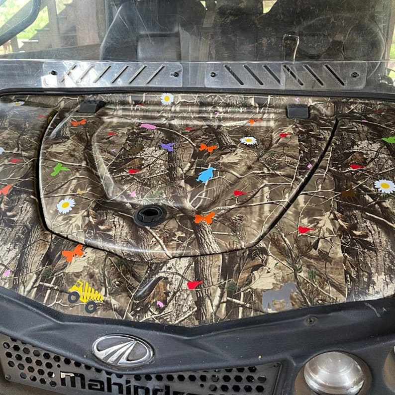 Puede incluir: Cap&oacute; de veh&iacute;culo utilitario Mahindra con un patr&oacute;n de camuflaje. El cap&oacute; est&aacute; decorado con mariposas de colores, margaritas y otros peque&ntilde;os gr&aacute;ficos. La rejilla frontal del veh&iacute;culo es negra con el logotipo de Mahindra.