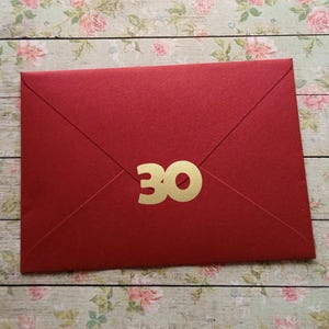 Könnte beinhalten: Ein roter Umschlag mit der goldenen Zahl 30. Der Umschlag ist verschlossen und hat eine klassische dreieckige Klappe. Der Hintergrund ist ein Blumenmuster auf einer Holzoberfläche.
