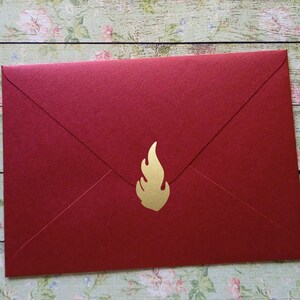 Op de afbeelding: Een rode envelop met een gouden vlamsticker. De envelop is gemaakt van getextureerd papier en is verzegeld. De gouden vlamsticker bevindt zich in de rechter benedenhoek.
