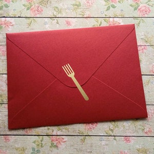 May include: A red envelope with a gold fork emblem. The envelope is closed and has a classic triangular flap. The background is a floral pattern on a light wood surface.