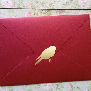 Pode incluir: Um envelope vermelho com uma silhueta dourada de um pássaro. O envelope está selado e tem uma aba triangular clássica. O pássaro, voltado para a esquerda, adiciona um toque decorativo à papelaria.
