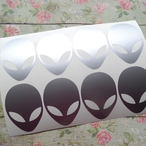 Könnte beinhalten: Bogen mit Aufklebern mit Alien-Kopf-Designs. Die Aufkleber sind in Silber und Dunkelgrau gehalten. Jeder Alien-Kopf hat große, mandelförmige Augen und ein glattes, ovales Gesicht. Die Aufkleber befinden sich auf einem weißen Hintergrund.
