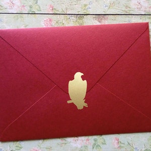May include: A red envelope with a gold eagle emblem. The envelope is rectangular with a pointed flap closure. The gold eagle is perched on a branch. The envelope is made of a textured paper.