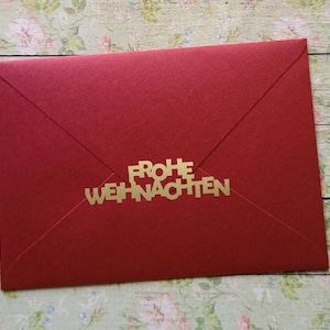 Op de afbeelding: Een rode envelop met de gouden tekst "FROHE WEIHNACHTEN". De envelop is verzegeld en heeft een klassieke driehoekige flap. De achtergrond is een bloemenpatroon.