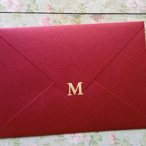Könnte beinhalten: Ein tiefroter Umschlag mit dem goldenen Buchstaben "M". Der Umschlag besteht aus strukturiertem Papier und hat ein klassisches, gefaltetes Design. Der Hintergrund ist eine helle Holzoberfläche mit floralen Mustern.