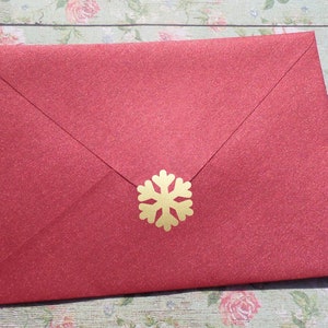 May include: A red glitter envelope with a gold snowflake embellishment. The envelope is closed and has a textured surface. The snowflake is centered on the front of the envelope.