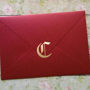 Könnte beinhalten: Ein roter Umschlag mit einem goldenen Buchstaben "C" auf der Vorderseite. Der Umschlag besteht aus strukturiertem Papier und ist versiegelt. Der Hintergrund ist ein Blumenmuster auf einer Holzoberfläche.
