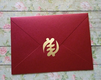 May include: A red envelope with a gold Adinkra symbol in the center. The envelope is made of a textured paper and has a classic envelope shape. The gold symbol is a stylized design.