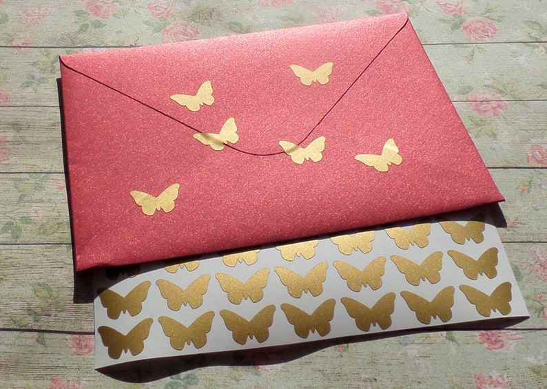 K&ouml;nnte beinhalten: Eine Blatt mit goldenen Schmetterlingsaufklebern auf wei&szlig;em Hintergrund. Das Blatt liegt auf einem roten Umschlag mit goldenen Schmetterlingsaufklebern.