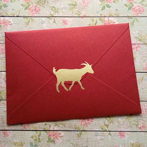 Op de afbeelding: Een rode envelop met een gouden geit silhouet. De envelop is verzegeld en heeft een eenvoudig, elegant ontwerp. De geit loopt naar rechts.