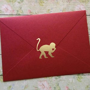 Puede incluir: Un sobre rectangular de color rojo que presenta una silueta dorada de un mono centrada. El sobre tiene un diseño clásico de solapa triangular y una textura mate y suave, creando un contraste marcado entre el papel rojo intenso y el icono del mono en oro.