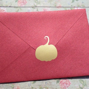 Puede incluir: Un sobre rojo con una pegatina dorada de calabaza. El sobre tiene un acabado texturizado y brillante. La pegatina de calabaza está centrada en la solapa del sobre, añadiendo un toque festivo. El fondo es un estampado floral.