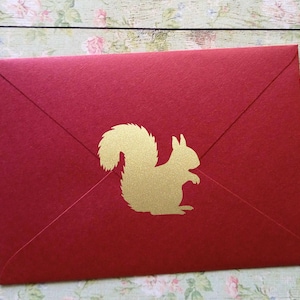 Pode incluir: Um envelope vermelho com uma silhueta dourada de esquilo. O envelope é feito de papel texturizado e tem uma aba triangular clássica. O esquilo está centrado na frente do envelope.