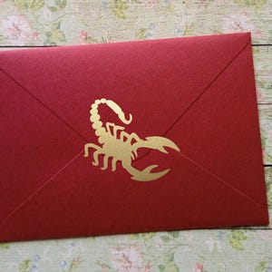 Könnte beinhalten: Ein roter Umschlag mit einem goldenen Skorpion-Design. Der Umschlag hat eine tiefrote Farbe und zeigt ein goldenes Skorpion-Emblem in der Mitte. Der Umschlag ist geschlossen und bereit zum Versiegeln.