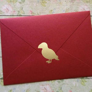 Könnte beinhalten: Ein roter Umschlag mit einem goldenen Vogelaufkleber. Der Umschlag ist geschlossen und hat eine dreieckige Klappe. Der Vogelaufkleber ist eine einfache Silhouette eines Vogels.