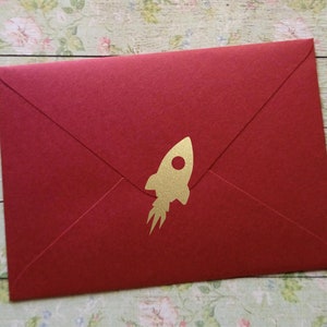 以下が含まれることがあります： 金色のロケットのデザインが施された赤い封筒。封筒は閉じられており、シンプルでエレガントなデザインです。ロケットは炎の軌跡を持つシルエットです。