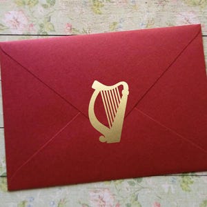 Könnte beinhalten: Ein roter Umschlag mit einem goldenen Harfenemblem. Der Umschlag besteht aus strukturiertem Papier und ist versiegelt. Das Harfenemblem ist ein traditionelles irisches Symbol, geeignet für Einladungen oder Ankündigungen.