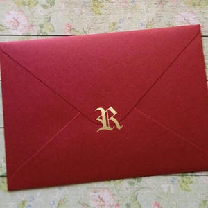 Może przedstawiać: Czerwona koperta ze złotą literą "R" pośrodku. Koperta wykonana jest z fakturowanego papieru i jest zapieczętowana. Tło to wzór kwiatowy.