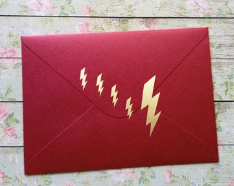 May include: A red envelope with a gold lightning bolt design. The envelope is rectangular and has a textured surface. The lightning bolts are arranged in a diagonal line across the front of the envelope.