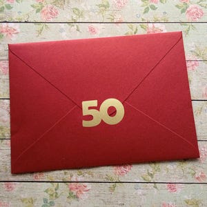 Könnte beinhalten: Ein roter Umschlag mit einer goldenen "50" auf der Vorderseite. Der Umschlag ist geschlossen und hat ein klassisches, gefaltetes Design. Der Hintergrund ist ein Blumenmuster auf einer Holzoberfläche.