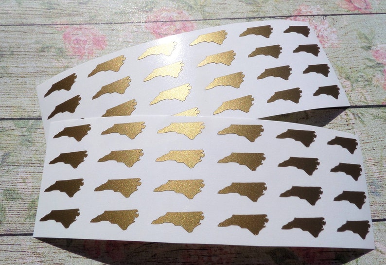 May include: Two sheets of white paper with multiple gold North Carolina state outline stickers. The stickers are arranged in rows.