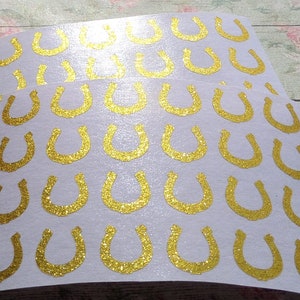 Può includere: Fogli di adesivi con forme di ferro di cavallo glitterate dorate. I ferri di cavallo sono disposti in file su sfondo bianco. Gli adesivi hanno una texture glitterata.