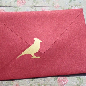 May include: A red envelope with a gold bird silhouette. The envelope has a textured, glittery finish. The bird is a simple, elegant design, adding a touch of sophistication to the stationery.