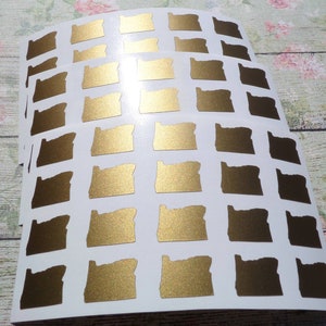 May include: A sheet of gold foil stickers featuring the outline of the state of Oregon. The stickers are arranged in a grid pattern on a white background.