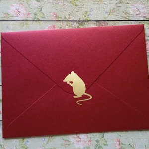 May include: A red envelope with a gold-colored mouse silhouette on the front. The envelope is sealed and has a classic triangular flap. The background is a floral pattern on a wooden surface.