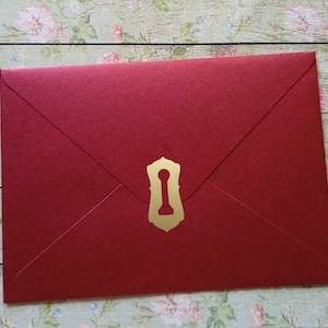 May include: A red envelope with a gold keyhole-shaped clasp. The envelope is rectangular with a pointed flap and is made of a textured paper. The gold clasp is centered on the front of the envelope.