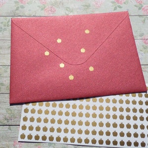 Può includere: Una busta rossa glitterata con dettagli di mele dorate e un foglio di adesivi a forma di mela dorata. La busta è chiusa e presenta piccoli disegni di mele dorate. Gli adesivi sono su sfondo bianco.