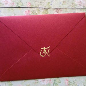 Pode incluir: Um envelope retangular vermelho escuro com acabamento em papel texturizado e aba pontiaguda. Centralizado na aba, há um símbolo Om em dourado metálico, um caractere sagrado das tradições orientais, aplicado como um selo decorativo.