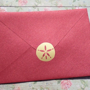 May include: Red envelope with a gold sand dollar sticker. The envelope is made of textured paper.