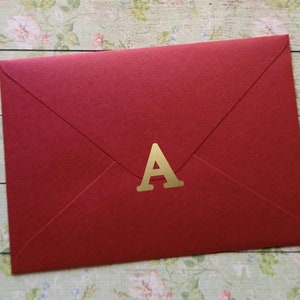 May include: A crimson envelope featuring a gold letter "A". The envelope is rectangular with a traditional flap design. The background displays a floral pattern.