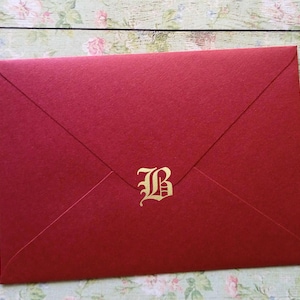May include: A red envelope with a gold letter "B" in a decorative font. The envelope is made of textured paper and has a classic, folded design. The background is a floral pattern.