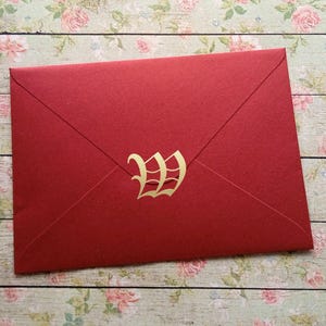 Peut inclure: Une enveloppe rouge avec un "W" doré sur le devant. L'enveloppe est scellée et présente un design classique. Le fond est un motif floral sur une surface en bois. Idéale pour les invitations ou les annonces. Mesure environ 16 cm.