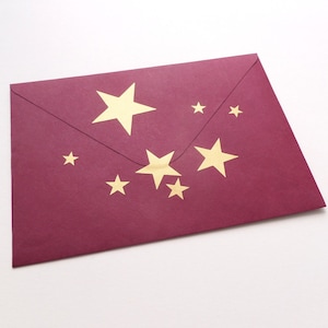 Può includere: Una busta color bordeaux con adesivi a forma di stella dorata. La busta è chiusa e pronta per essere spedita.