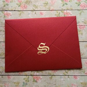 Könnte beinhalten: Ein roter Umschlag mit einem goldenen Buchstaben "S" auf der Vorderseite. Der Umschlag besteht aus strukturiertem Papier und ist versiegelt. Der Hintergrund ist ein Blumenmuster auf einer Holzoberfläche.