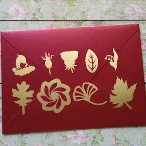 Pode incluir: Um envelope vermelho escuro com ilustrações botânicas douradas. As ilustrações incluem várias folhas, flores e outros elementos vegetais. O envelope está selado e pronto para uso.