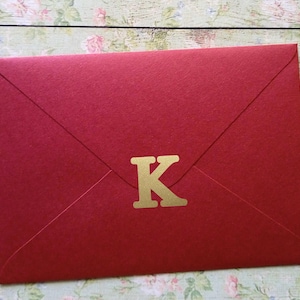 Könnte beinhalten: Ein roter Umschlag mit einem goldenen "K"-Buchstabenaufkleber. Der Umschlag besteht aus strukturiertem Papier und ist versiegelt. Der Hintergrund ist ein Blumenmuster auf einer Holzoberfläche.