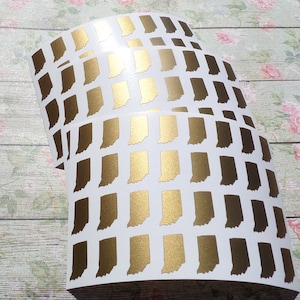 May include: Sheets of white stickers with a gold metallic outline of the state of Indiana. The stickers are arranged in a grid pattern, with multiple sheets visible. The stickers have a shiny, reflective surface.