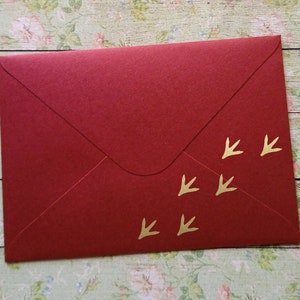 Op de afbeelding: Een dieprode envelop met een gouden vogelvoetafdrukontwerp. De envelop is gemaakt van getextureerd papier en heeft een klassieke klepsluiting. De gouden voetafdrukken zijn diagonaal over de voorkant geplaatst.